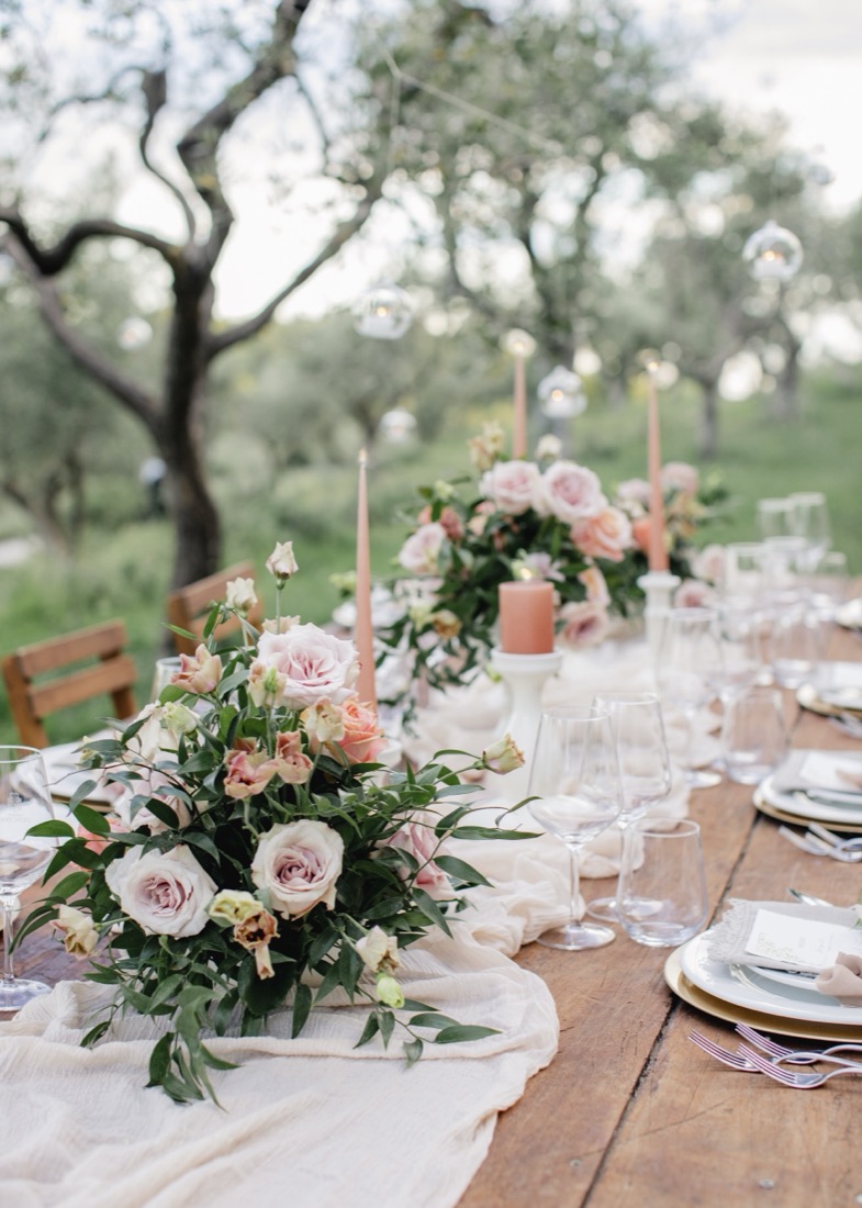 Reception table set with blush and peach roses, taper candles and hanging lanterns among olive trees