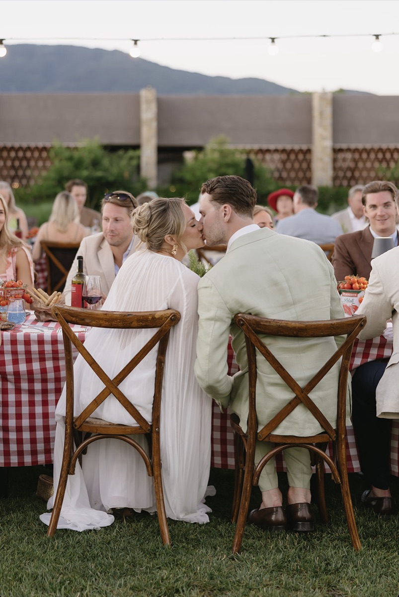 Couple kissing at a long picnic-style dinner table with red-and-white checkered tablecloths