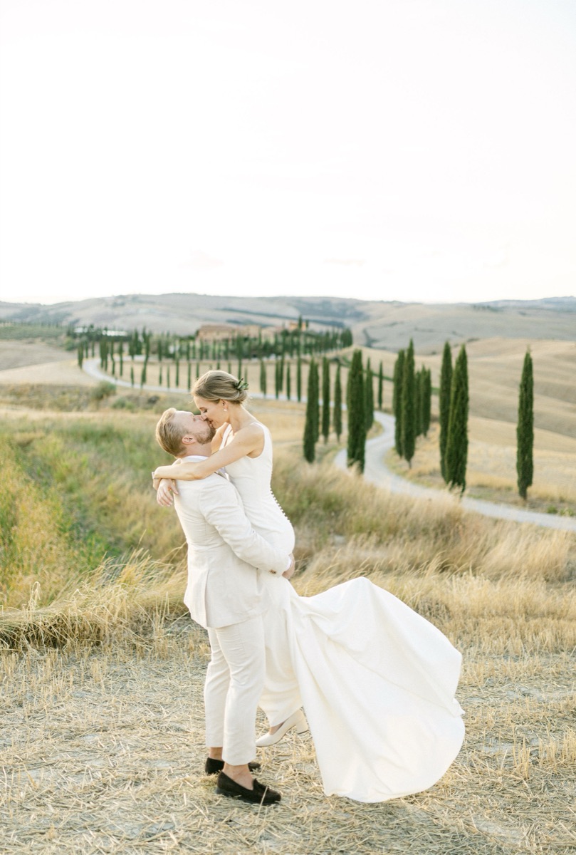 Groom lifting and kissing bride on a hillside with a cypress-lined Tuscan road behind them