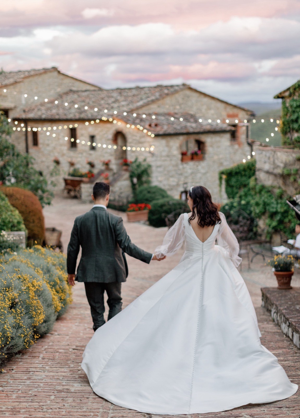 Bride and groom walking hand in hand toward a string-lit stone farmhouse at sunset