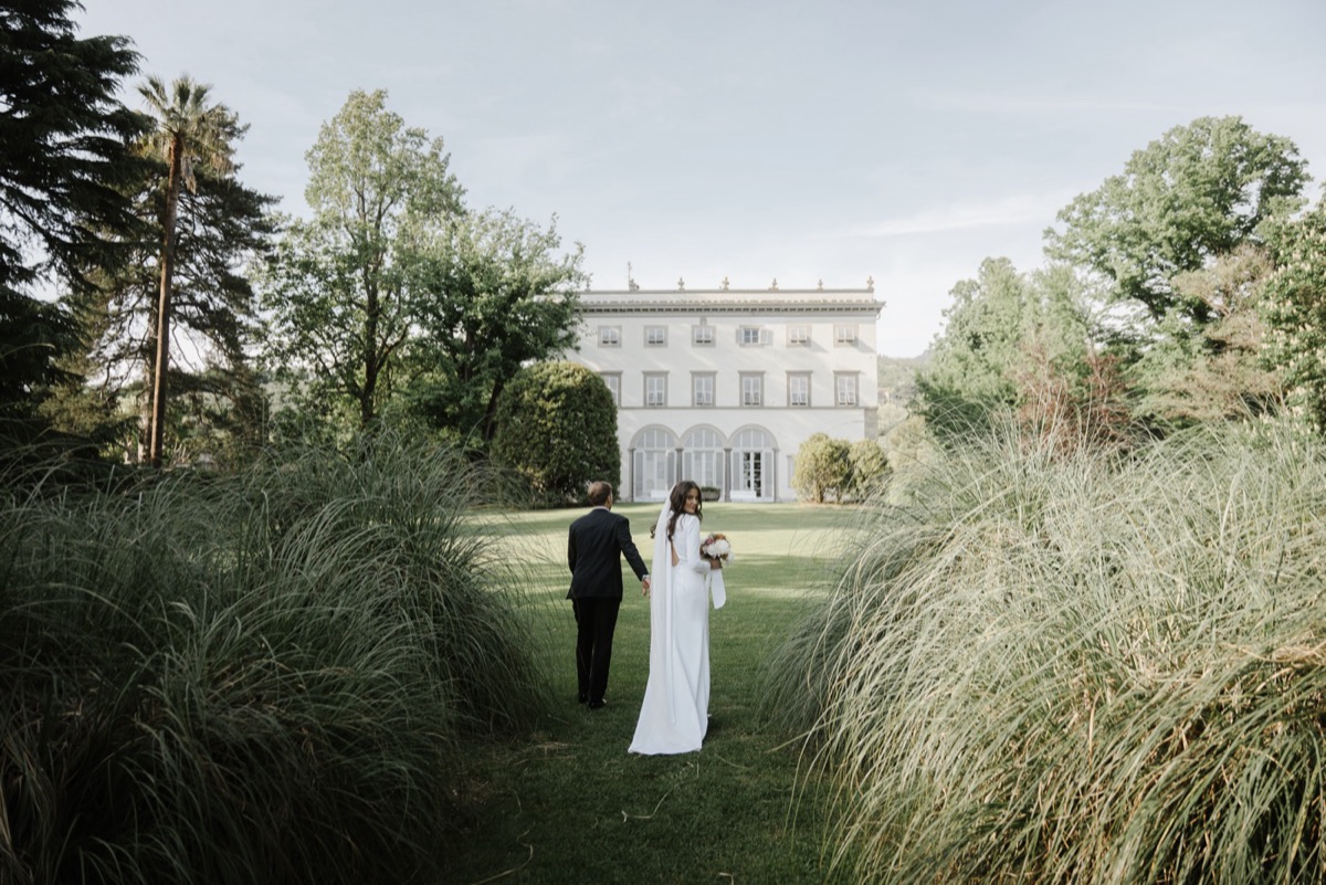 Bride and groom walking hand in hand across the lawn toward a grand Italian villa