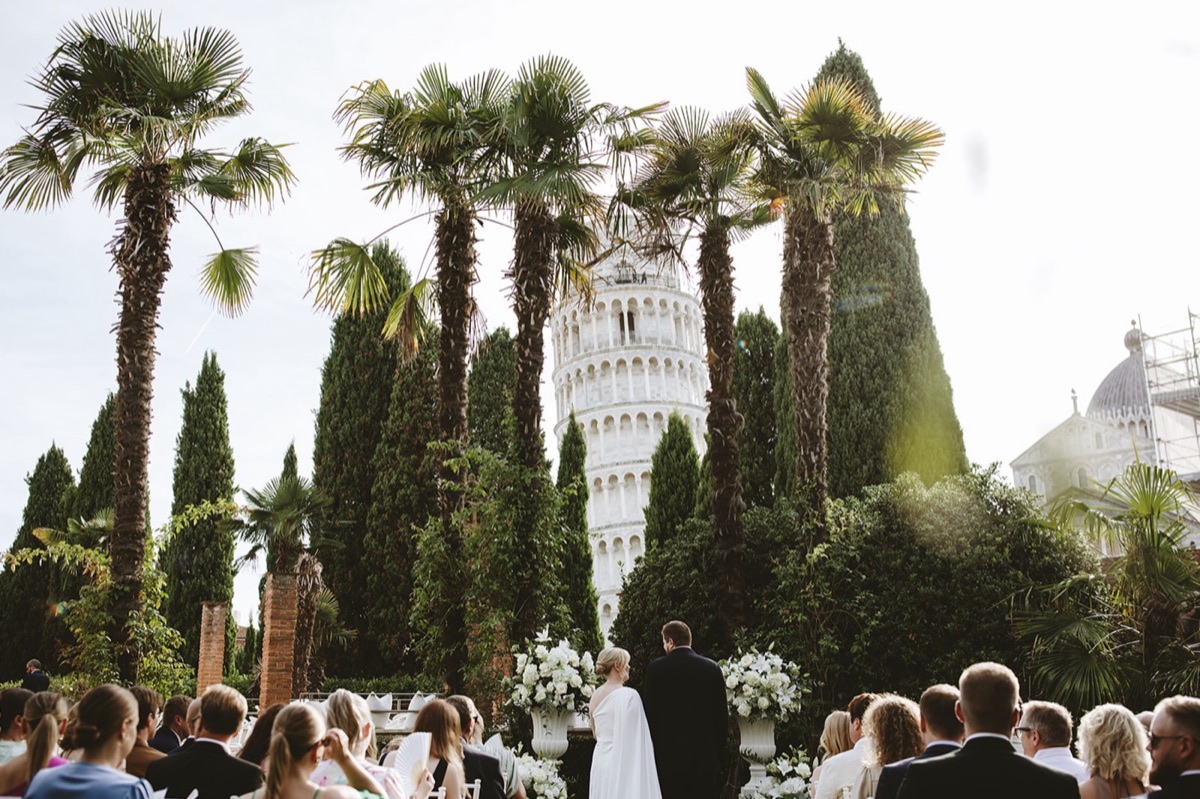 Wedding ceremony framed by palm trees and cypress with the Leaning Tower of Pisa rising behind
