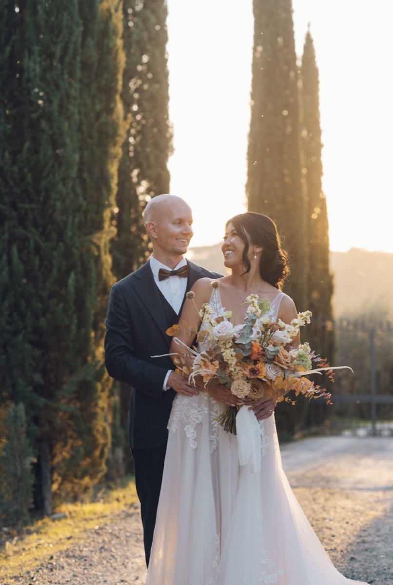 Lai and Thomas smiling at each other beside tall cypress trees, holding an autumnal bouquet