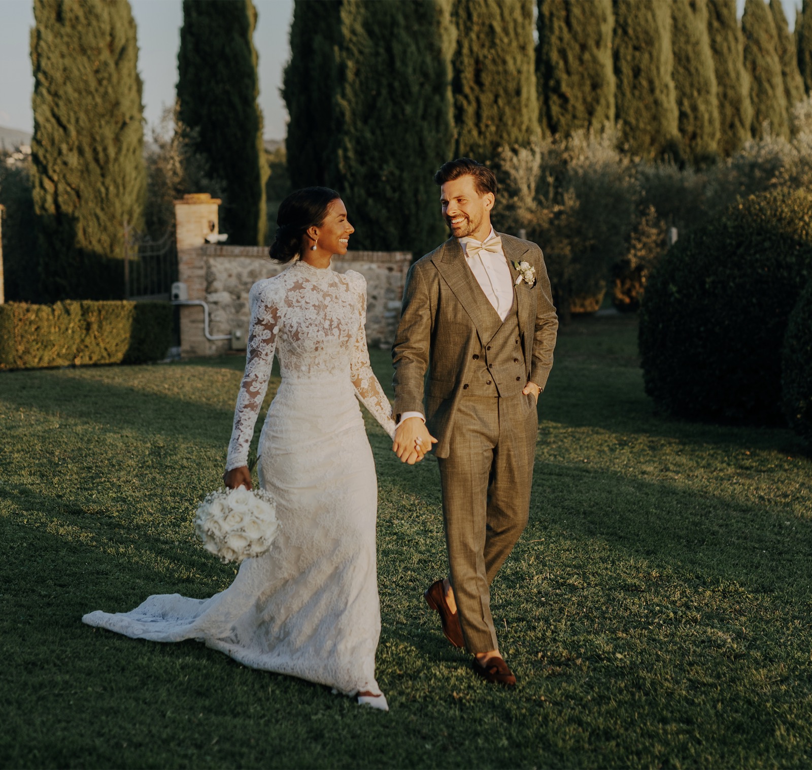 Isatou and Sami walking hand in hand through a cypress-lined Tuscan garden
