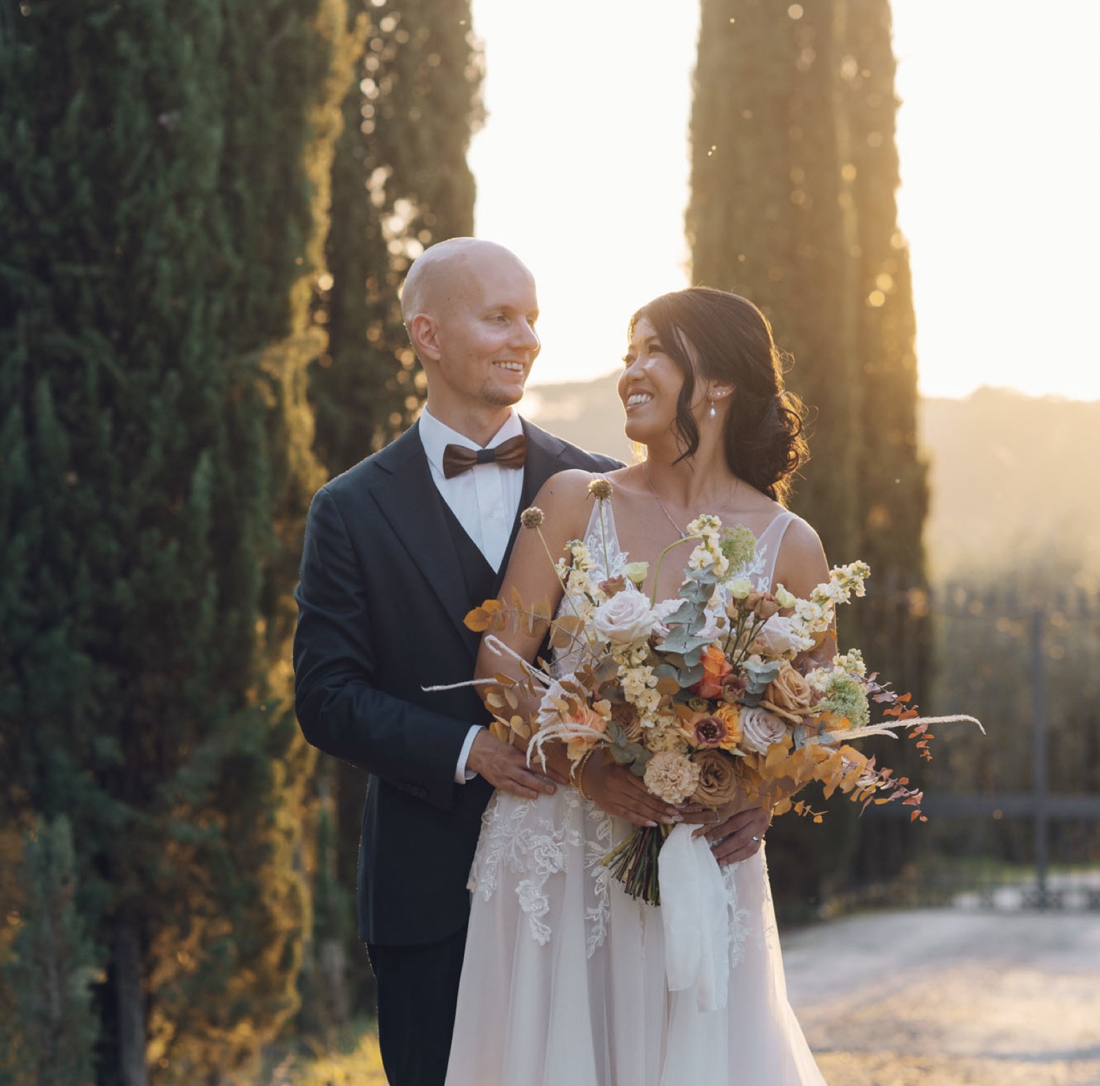 Lai and Thomas smiling at each other beside tall cypress trees, holding an autumnal bouquet