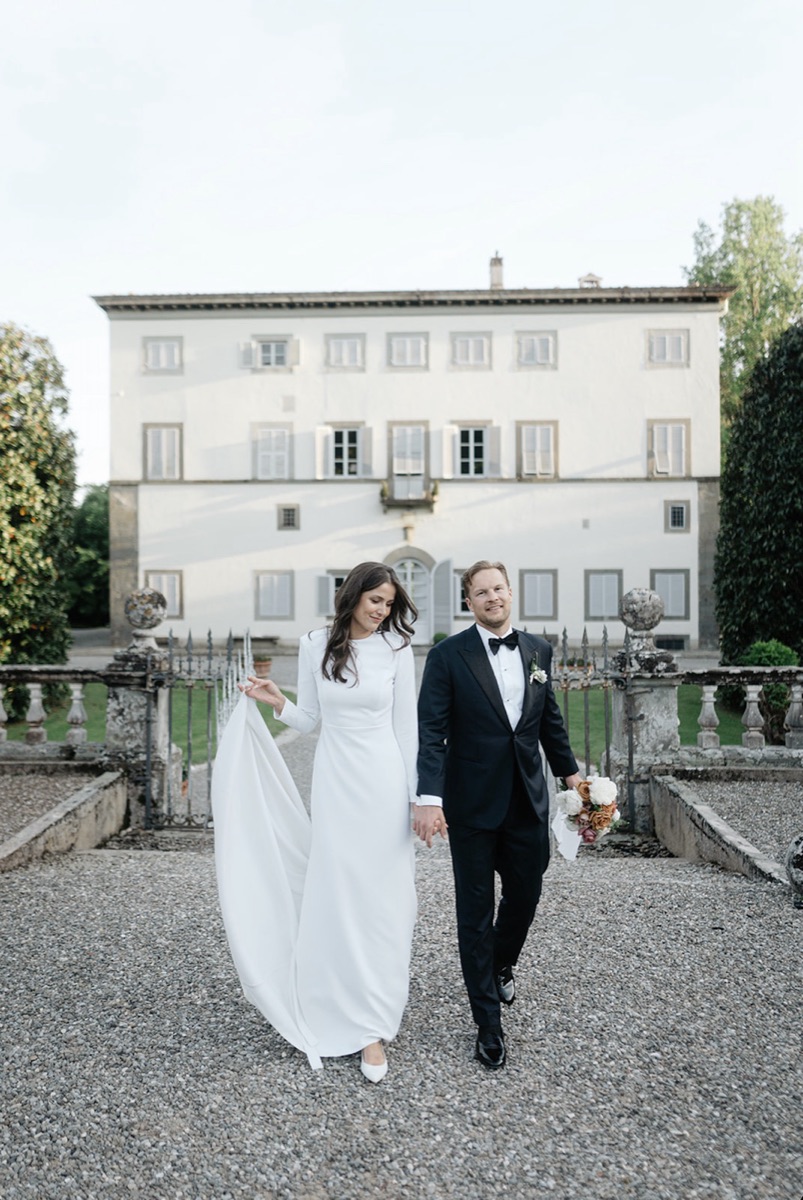 Emma and Teemu walking hand in hand up the gravel path toward a Tuscan villa